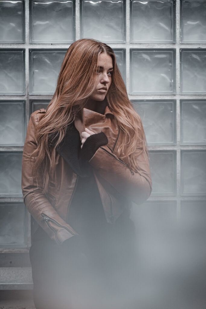Frau mit langen rötlichen Haaren in brauner Jacke vor einer Glasbausteinwand