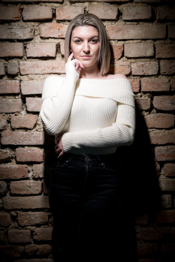 Frau im weißen Pullover lehnt an einer Backsteinwand, Hand am Kinn