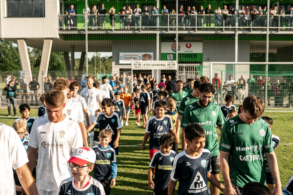 Fußballspieler laufen vor dem Spiel auf den Platz, auf der Tribüne sitzen Zuschauer