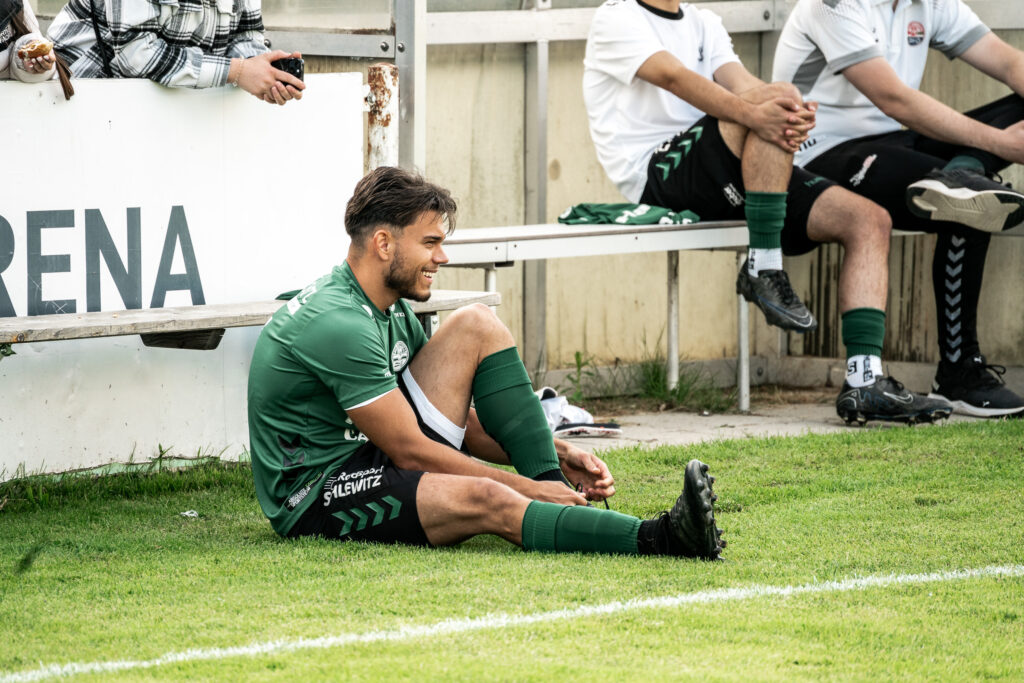 Fußballspieler in grünem Trikot sitzt am Spielfeldrand auf dem Rasen